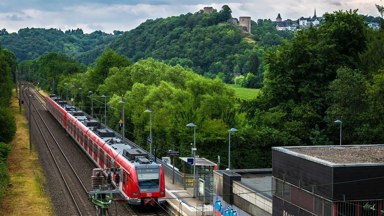 Regionalzug an einem begrünten Bahnsteig vor einer Anhöhe mit Burgruine